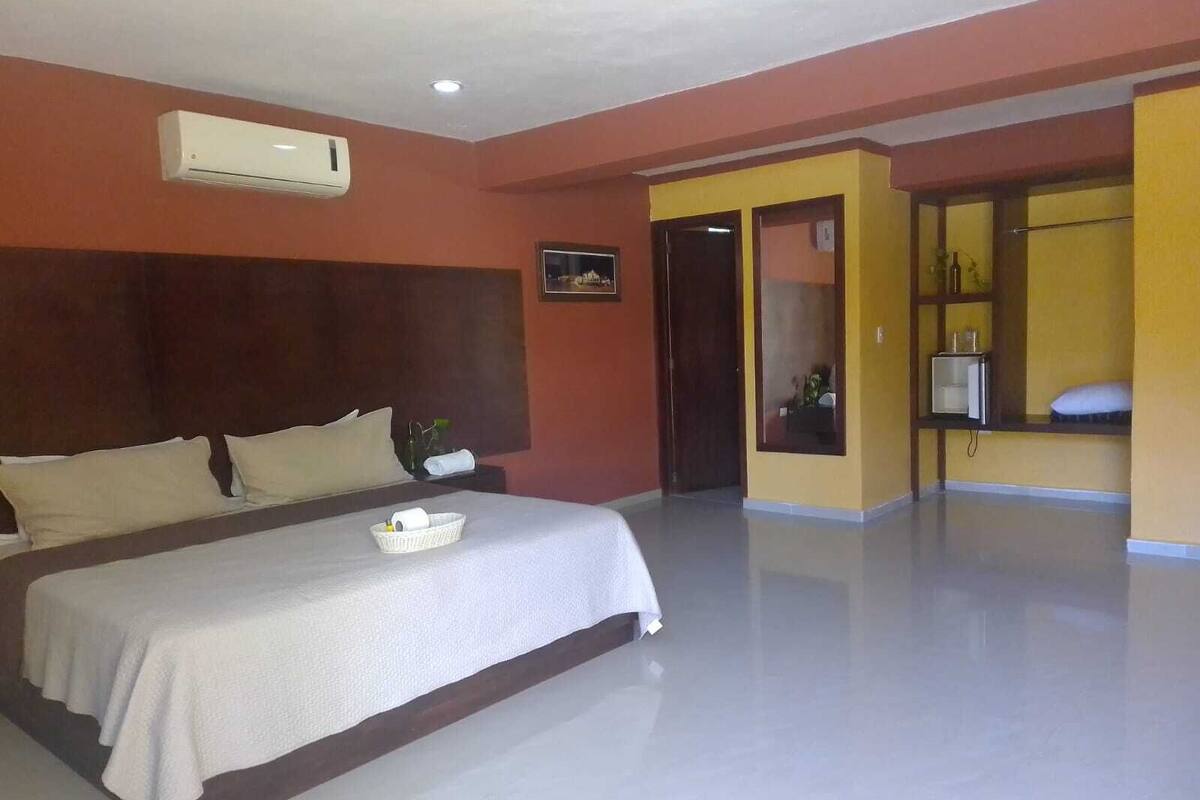 A hotel room with a large bed, a nightstand, an air conditioning unit, a door leading to another room, and a small shelf with items.