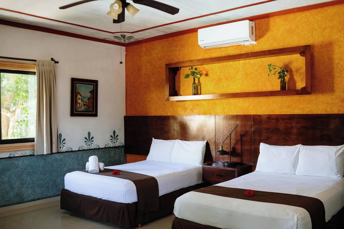 A hotel room with two beds, a window with curtains, a painting, an air conditioning unit, and a shelf with a plant.