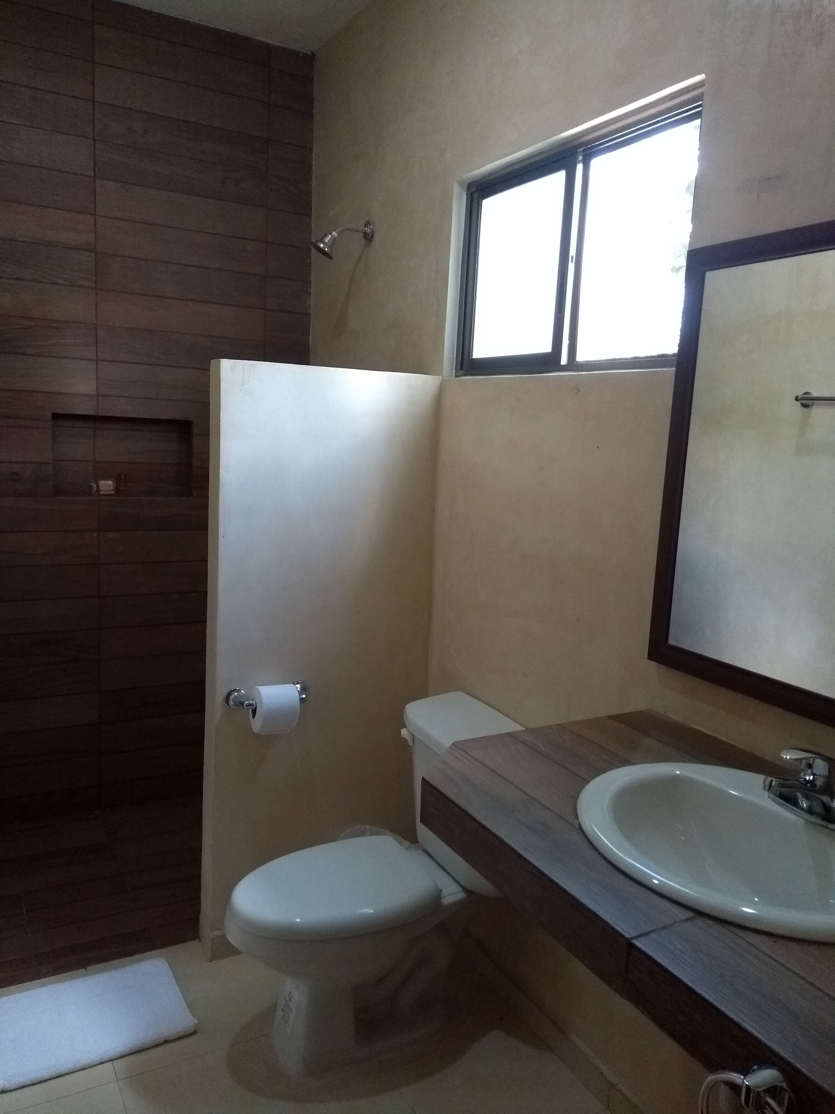 A bathroom with a wooden shower enclosure, a toilet, a sink with a mirror, and a window providing natural light.