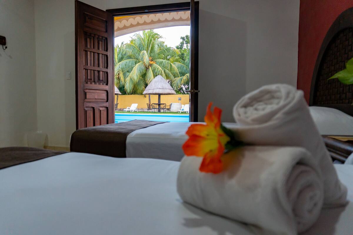 A hotel room with a bed, a rolled towel with a flower, and a view of a pool and palm trees through an open door.