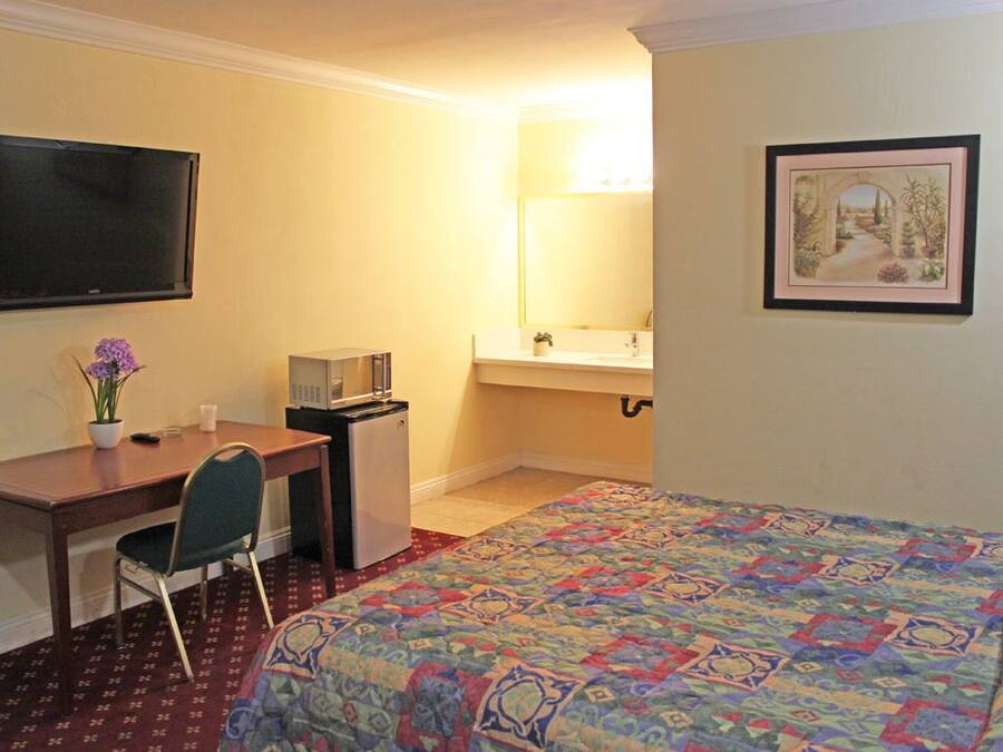 Habitación de hotel con cama, escritorio con silla, televisor, refrigerador pequeño, lavamanos y un cuadro enmarcado en la pared.