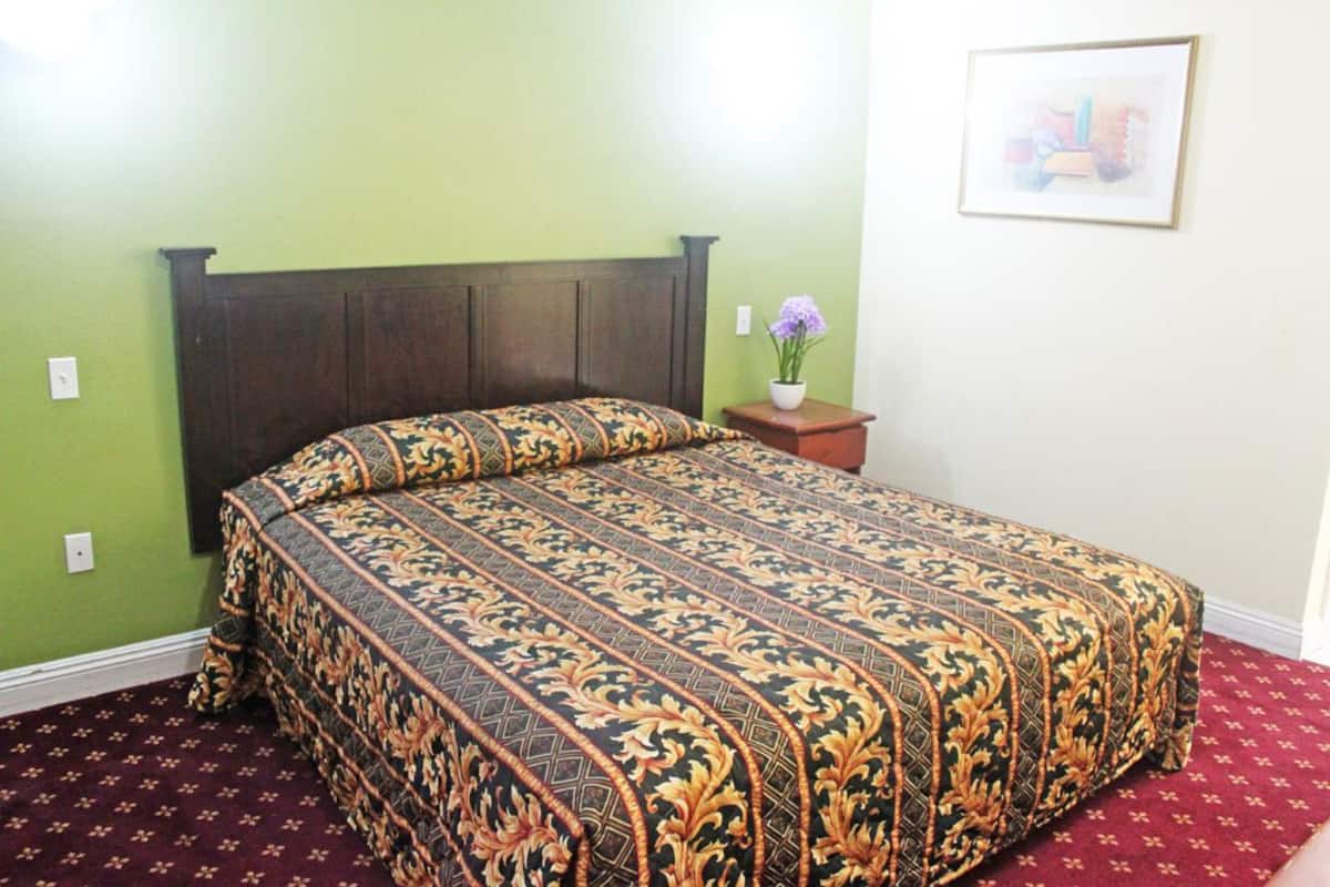 A bedroom with a dark wooden headboard, a patterned bedspread, a small bedside table with a vase of flowers, and a framed picture on the wall.