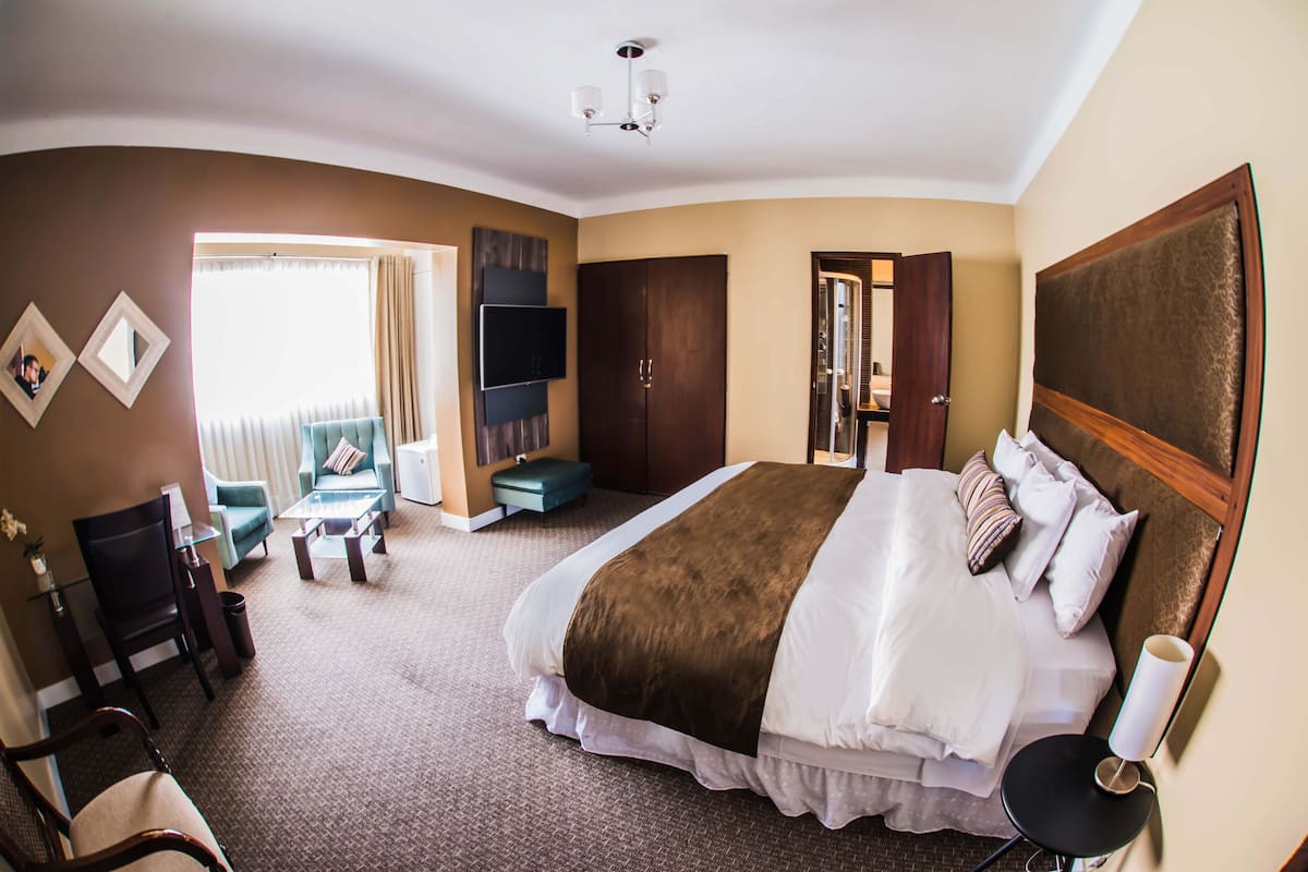 Ein Hotelzimmer mit einem großen Bett, einem an der Wand befestigten Fernseher, einer Sitzecke mit einer Couch und Sesseln sowie einer Tür zu einem weiteren Raum.