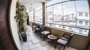 A balcony with a potted plant, wooden chairs, a small table, and a view of buildings through large windows. - Los Leones Hotel Boutique (Arequipa)