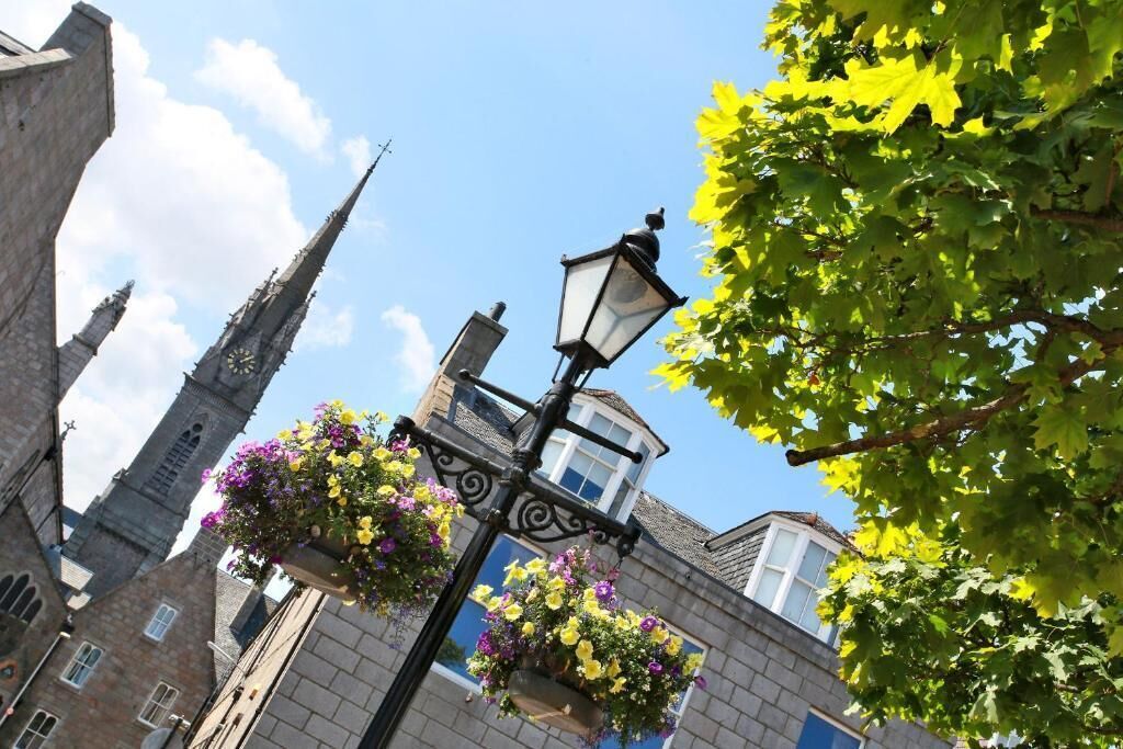 Un farol con cestas de flores colgantes y un edificio con ventana visible y una torre.