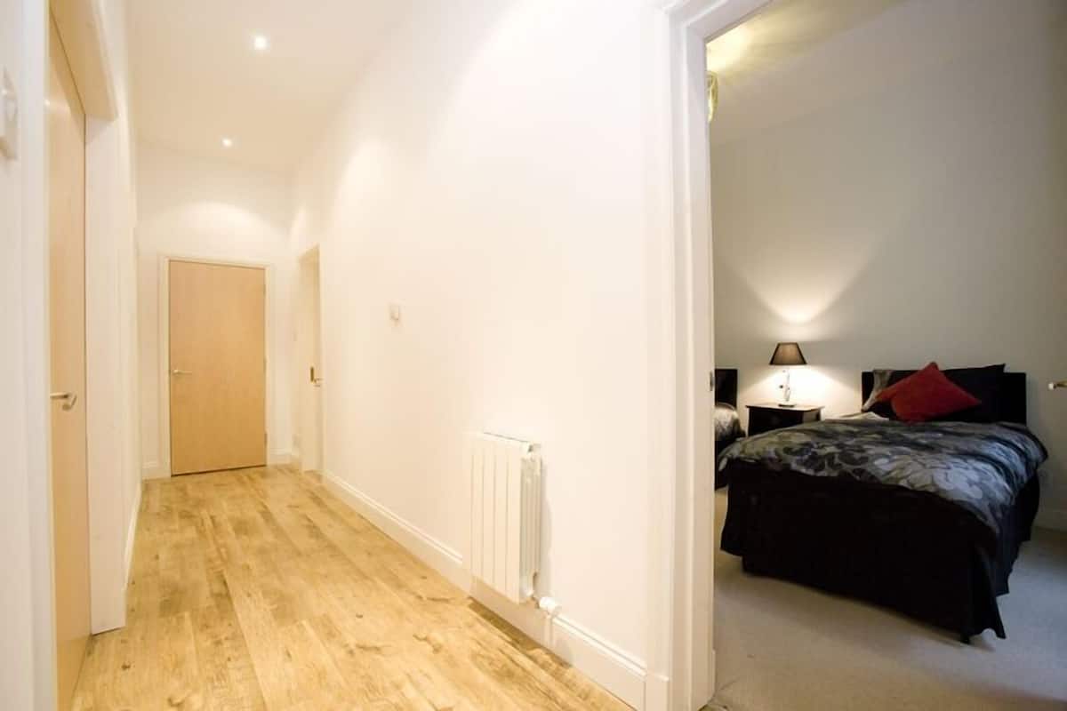 A narrow hallway with a wooden floor leading to a bedroom with a bed, a nightstand, and a lamp.