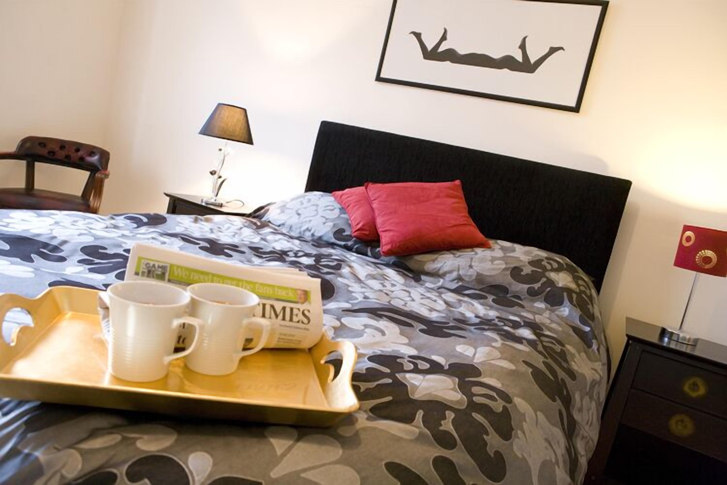 A neatly made bed with a patterned bedspread, a tray with two white mugs and a book, a lamp on a bedside table, and a framed picture on the wall.