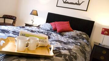 A neatly made bed with a patterned bedspread, a tray with two white mugs and a book, a lamp on a bedside table, and a framed picture on the wall.