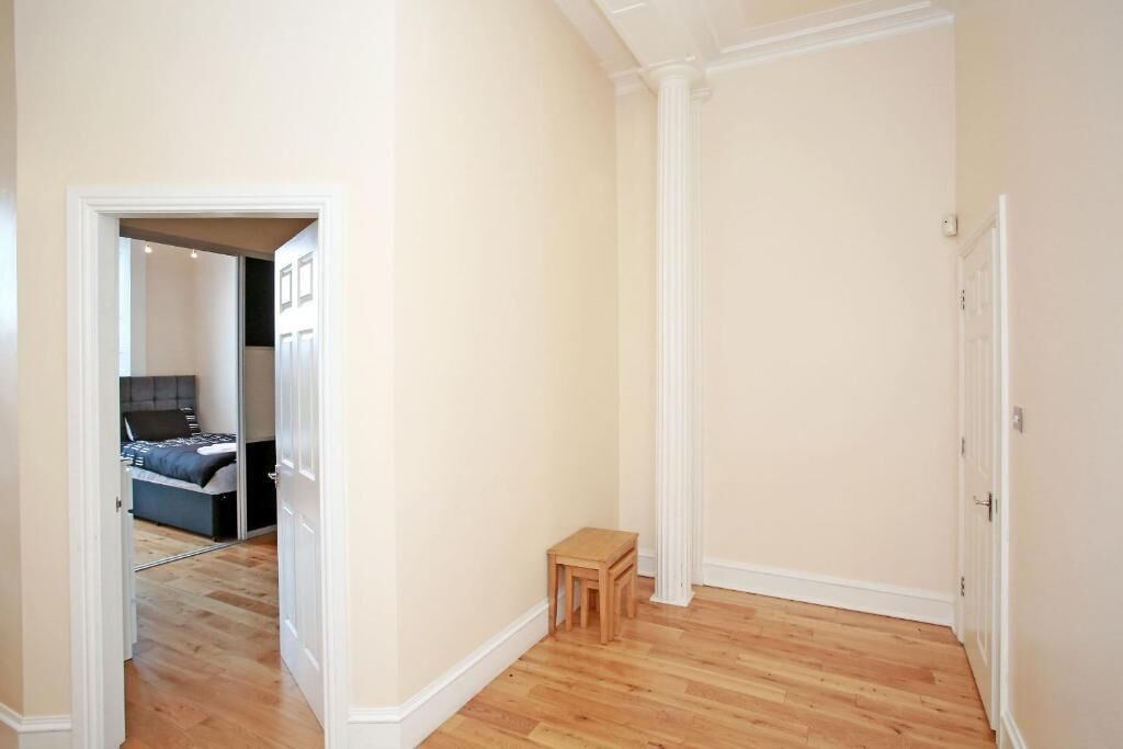 A hallway with wooden flooring, a small wooden stool, and a door leading to a bedroom with a bed and a nightstand.