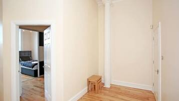 A hallway with wooden flooring, a small wooden stool, and a door leading to a bedroom with a bed and a nightstand.