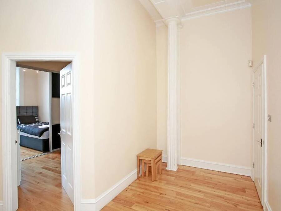A hallway with wooden flooring, a small wooden stool, and a door leading to a bedroom with a bed and a nightstand.