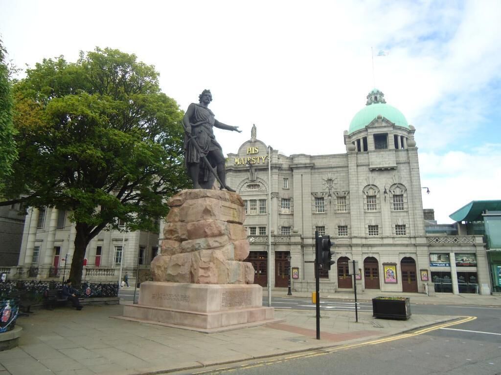 Eine Statue auf einem Sockel vor einem Gebäude mit einer Kuppel und der Inschrift 'HIS MAJESTY'.