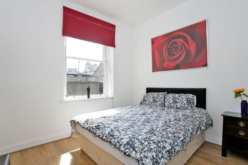 A bedroom with a bed, a window with a red blind, a wall-mounted rose painting, and a nightstand with a vase.