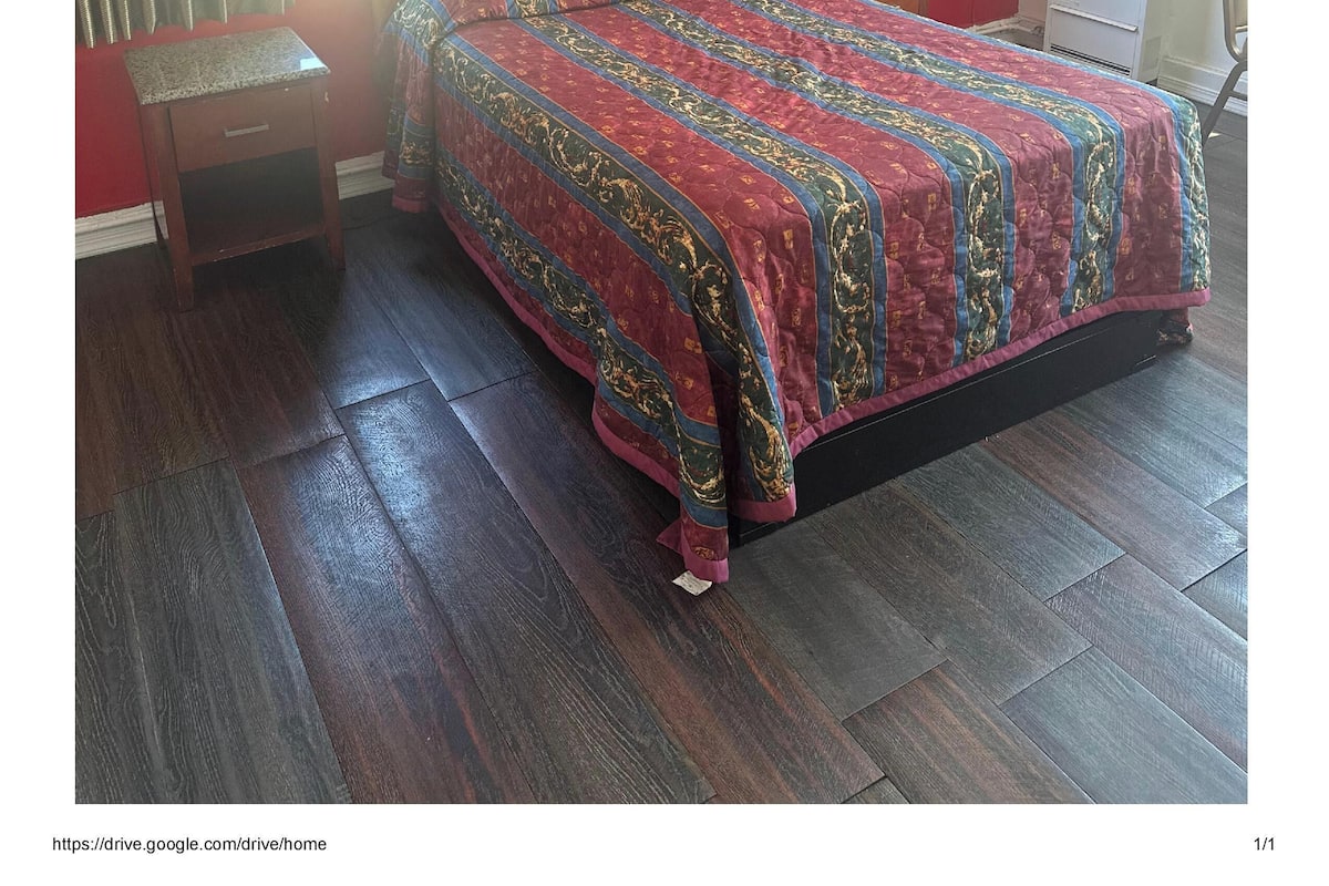Un dormitorio con cabecera de madera, cama con colcha estampada, mesita de noche, ventana con cortinas y radiador.
