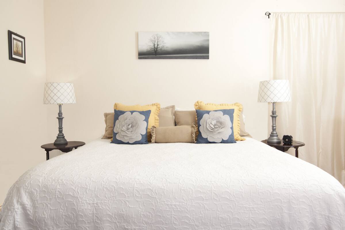 A neatly made bed with a white bedspread and decorative pillows, two bedside lamps, a framed picture on the wall, and a window with curtains.