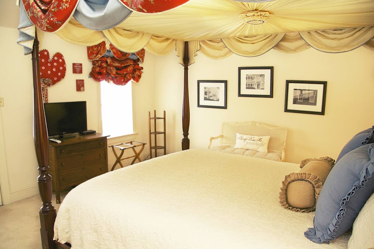 A bedroom with a large bed, a wooden dresser, a television on the dresser, a ladder, and three framed pictures on the wall.