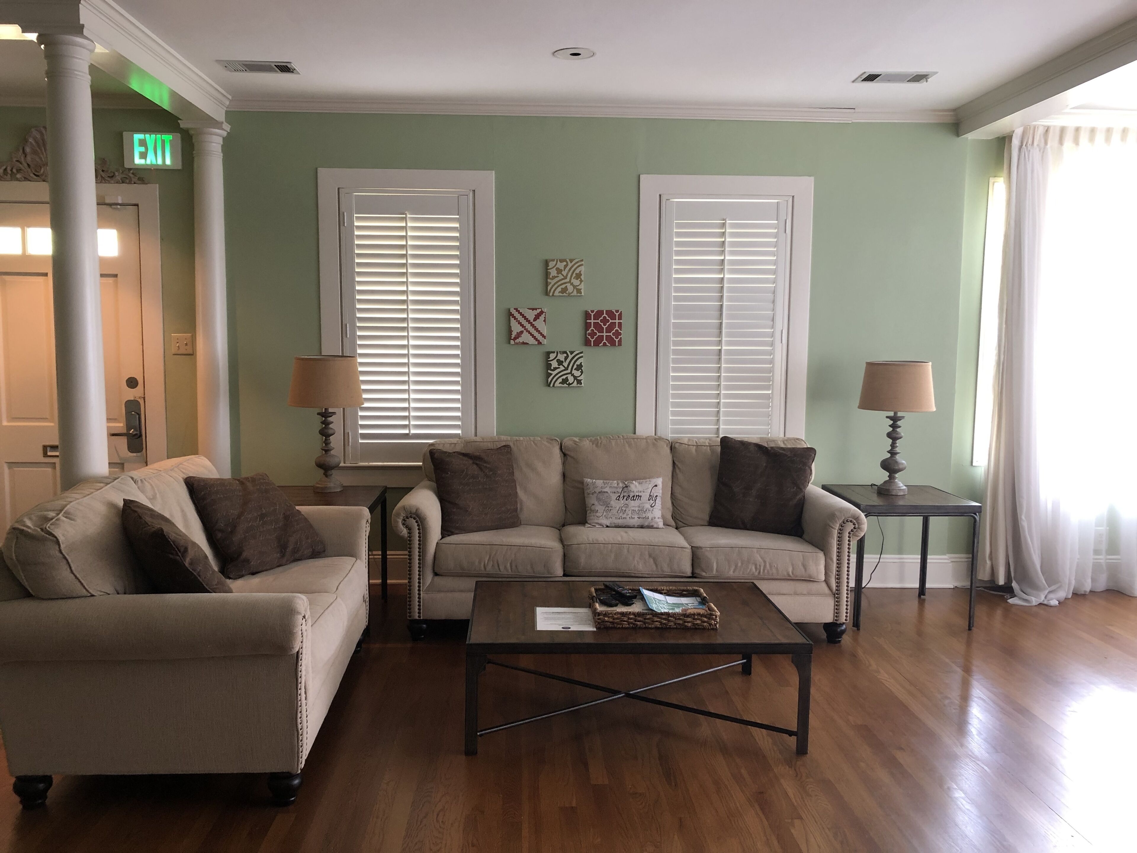 Sala de estar com dois sofás, uma mesa de centro e duas luminárias. Há duas janelas com persianas, uma parede com objetos decorativos e uma placa de saída.