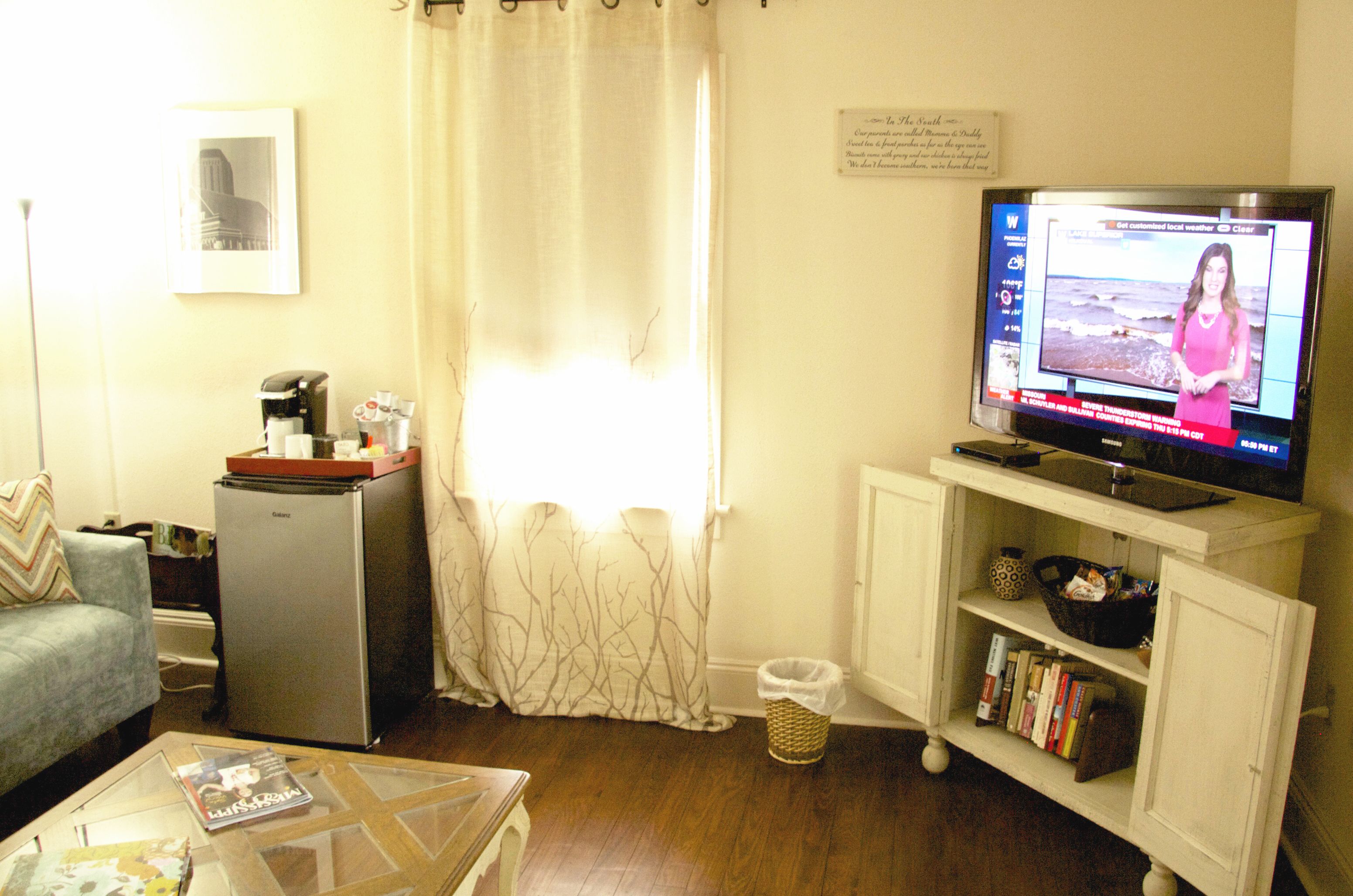 A living room with a television displaying a news channel, a refrigerator, a coffee maker, a bookshelf, and a window with a curtain.