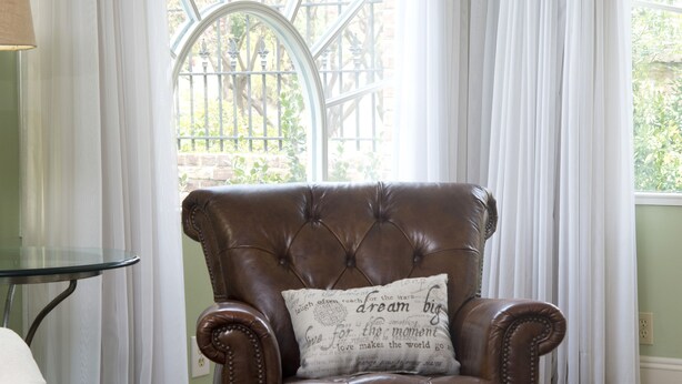 A brown leather armchair with a cushion that has text, positioned in front of a window with white curtains. - The Natchez Pearl (Natchez)