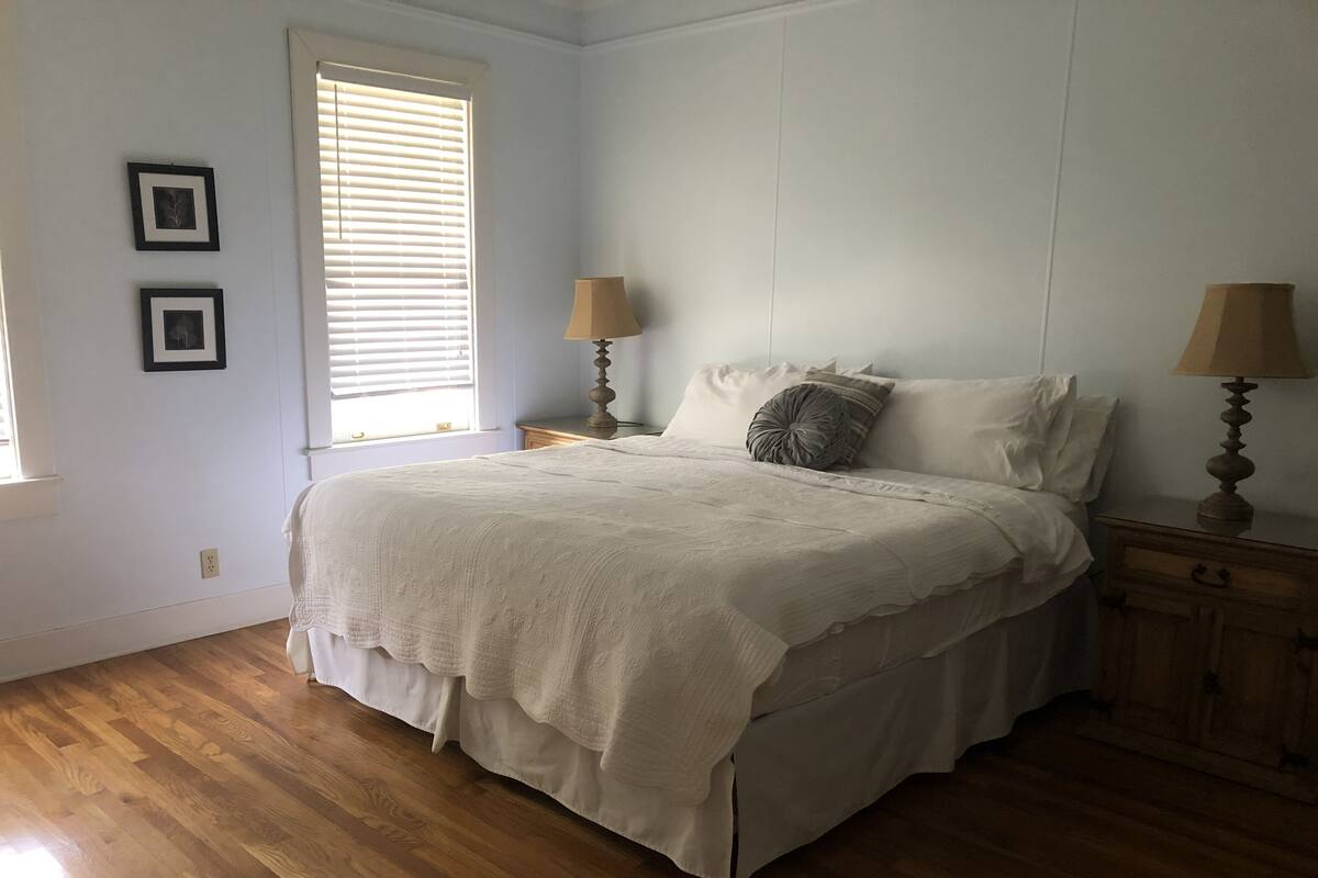 A bedroom with a large bed, two bedside tables with lamps, a window with blinds, and two framed pictures on the wall.