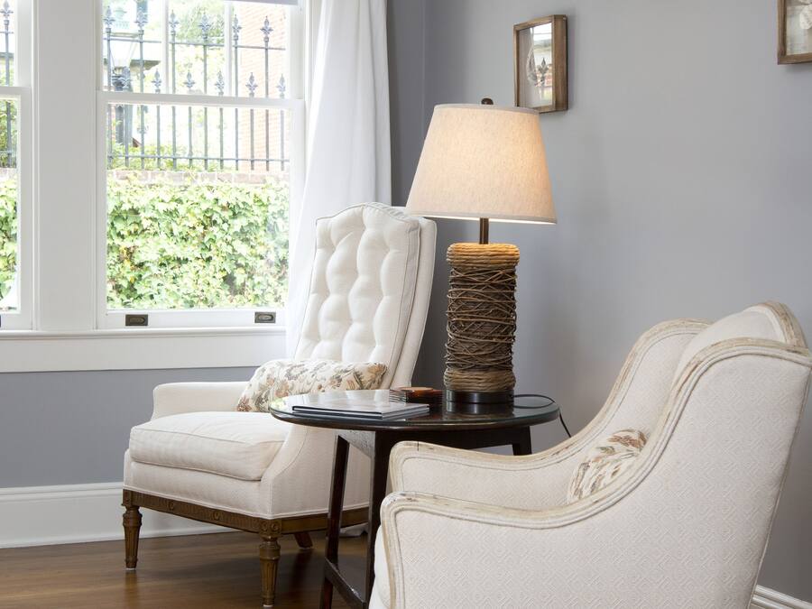 A room with two upholstered chairs, a small table with a lamp, and a window with a view of greenery outside.