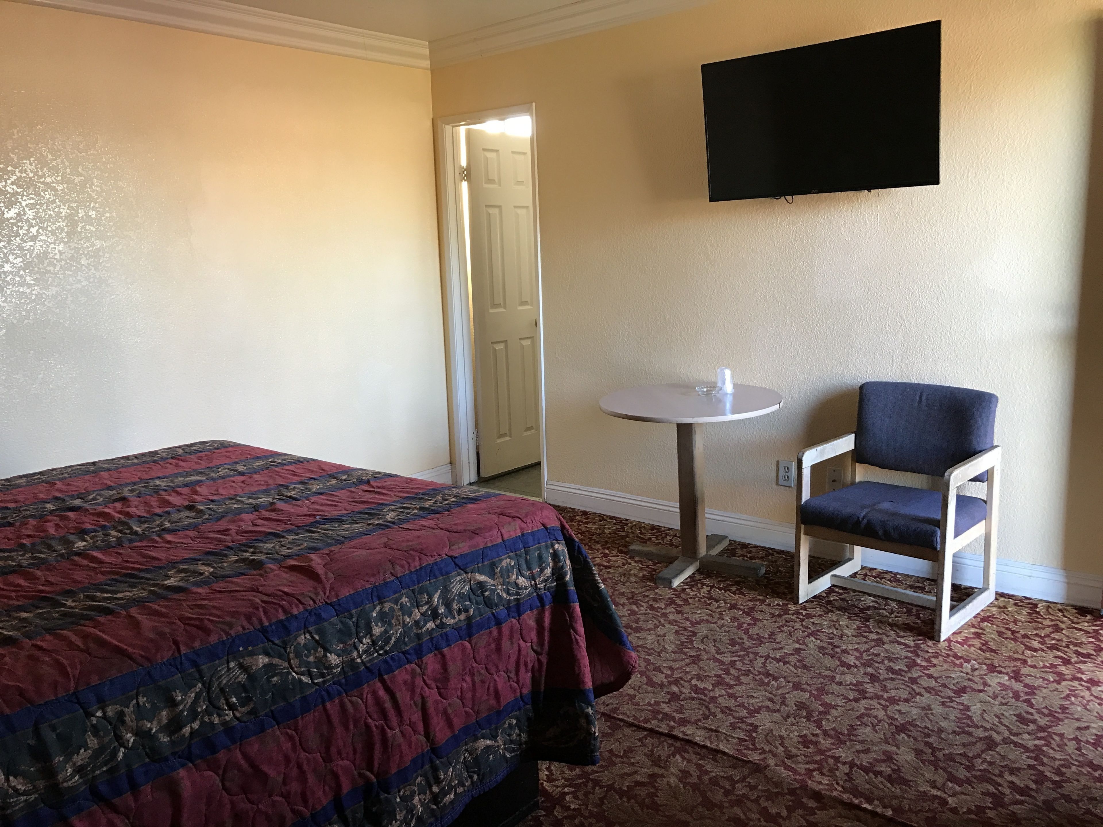 A hotel room with a bed, a small round table, a chair, and a flat-screen TV mounted on the wall.