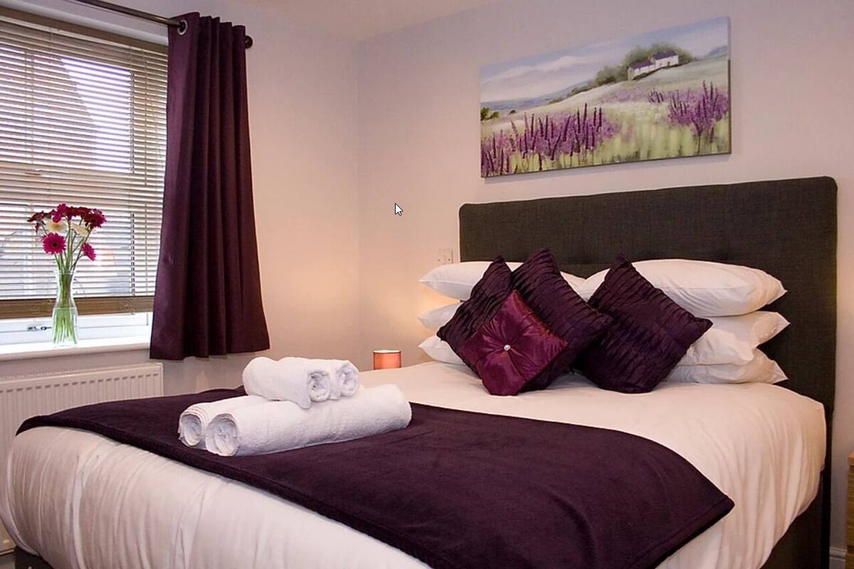 A neatly made bed with a dark headboard, white bedding, and purple pillows. A vase with flowers is on the windowsill, and a painting of a field is on the wall.