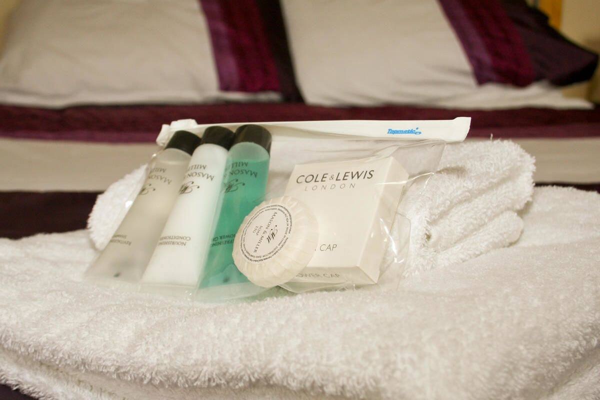 Toiletries including a bottle of lotion, a tube of cream, and a bottle of shampoo, along with a toiletry bag and a cap, on a white towel.