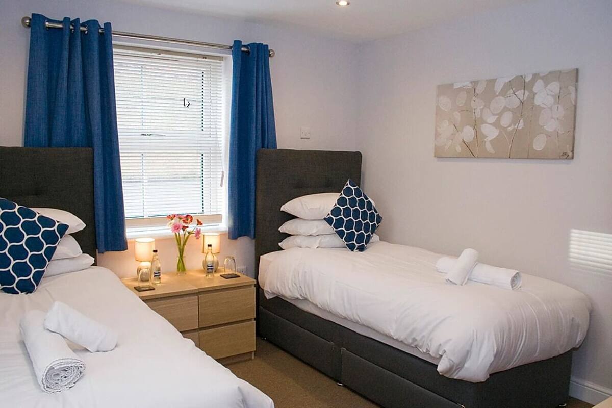 A hotel room with two single beds, a bedside table with a lamp, a vase with flowers, and a window with blue curtains.