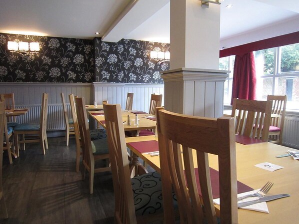 A dining area with wooden tables and chairs, a patterned wallpaper, and a window with a red curtain.