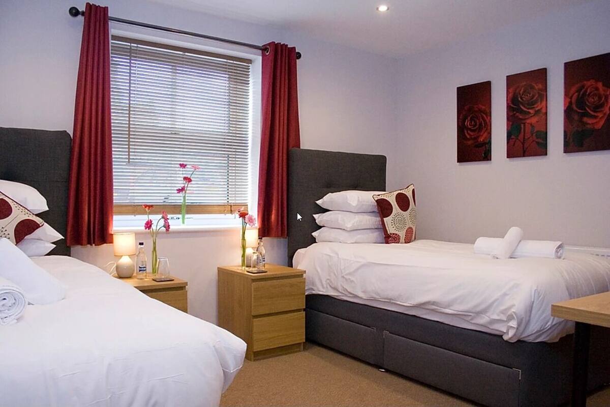 A hotel room with two beds, a window with red curtains, a bedside table with a lamp, and three rose paintings on the wall.