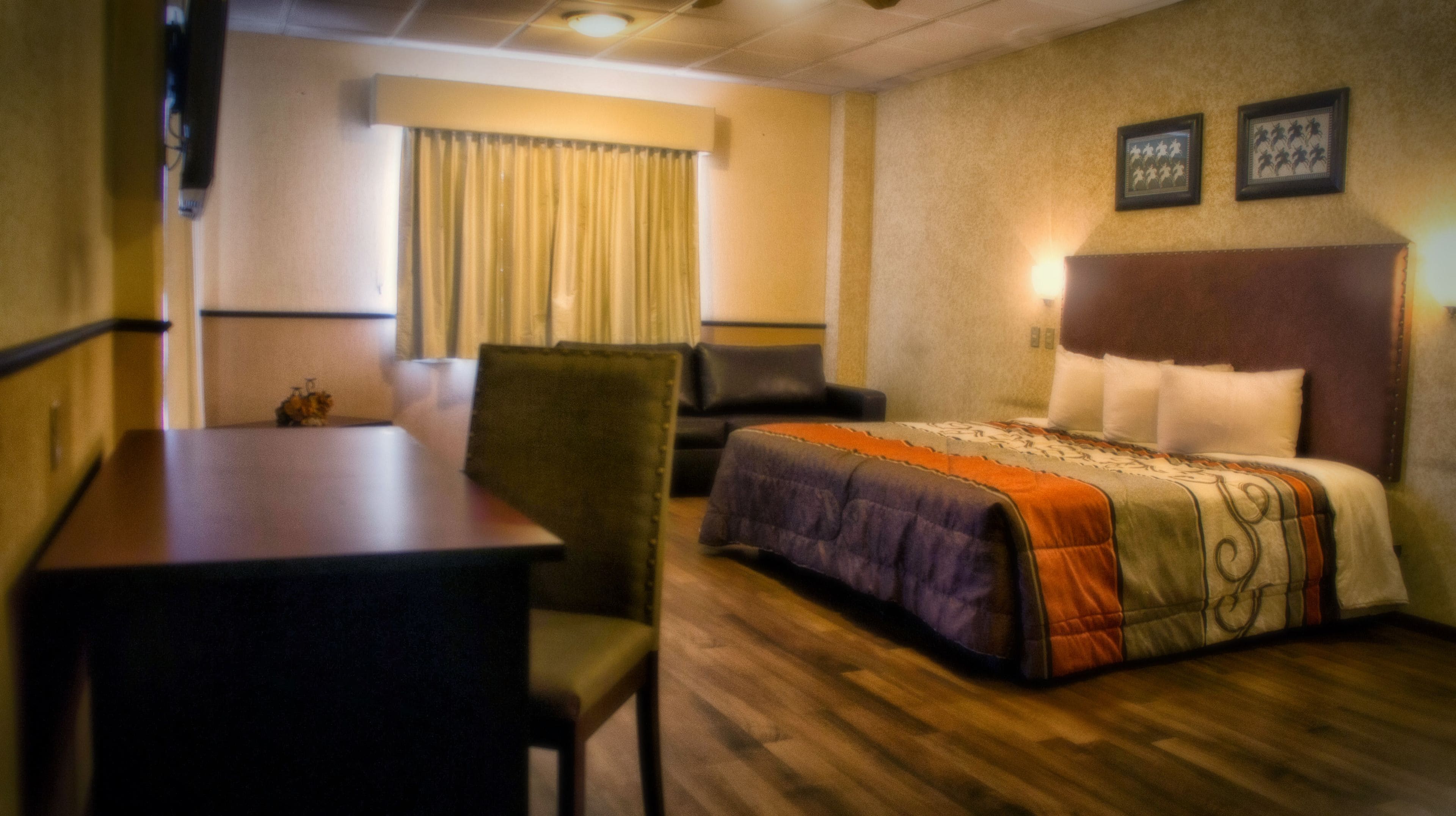 A hotel room with a bed, a desk, a chair, a window with curtains, and a wall with framed pictures.