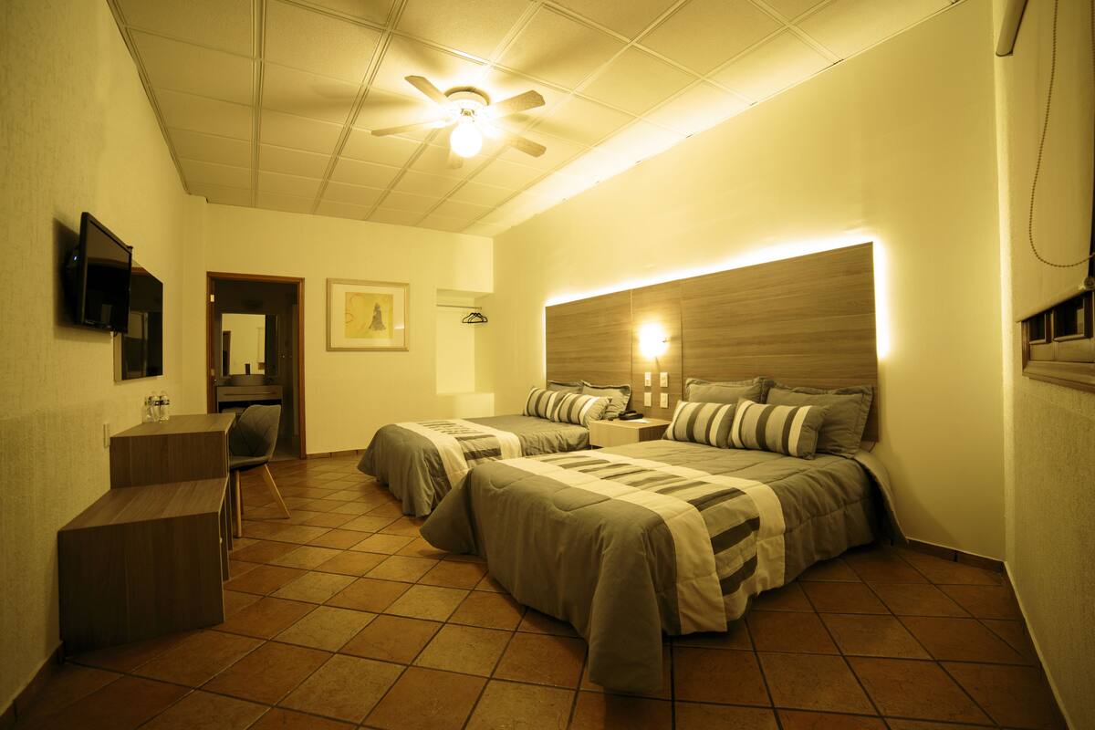 Habitación de hotel con dos camas, un escritorio, una silla, un ventilador de techo y un televisor montado en la pared.
