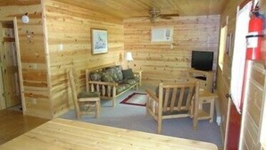 L’intérieur d’un chalet en bois, avec un canapé, des fauteuils, un téléviseur et un ventilateur de plafond.