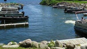 Une rivière bordée de bateaux amarrés, une jetée en bois et une zone herbeuse parsemée de rochers.