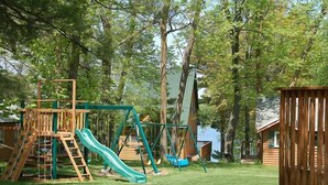 A playground with a slide and swings, surrounded by trees and a wooden cabin. - Woodland Beach Resort (Deerwood)