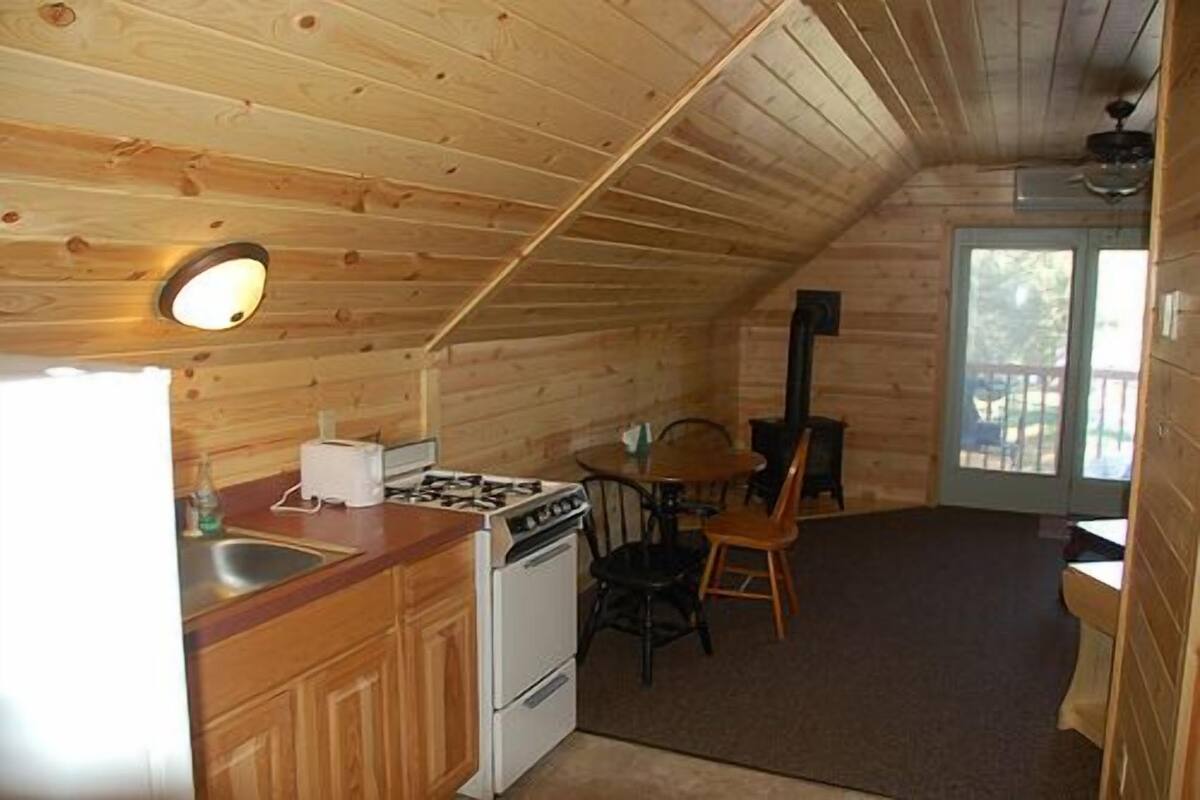 A small, wooden cabin with a kitchen area featuring a stove, sink, and cabinets. There is a dining table with chairs and a large window providing natural light.