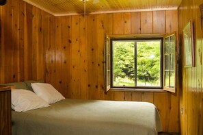 A wooden-paneled room with a bed, a window with a view of greenery, and a framed picture on the wall. - Woodland Beach Resort (Deerwood)
