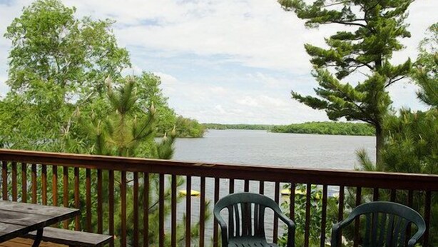 A wooden deck with two chairs and a table overlooking a lake, surrounded by trees. - Woodland Beach Resort (Deerwood)