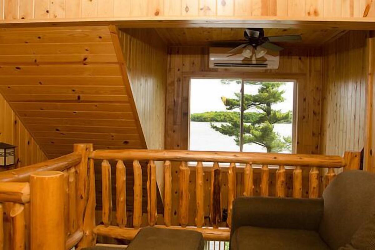 A wooden cabin with a balcony, a ceiling fan, and a view of a tree through a window.