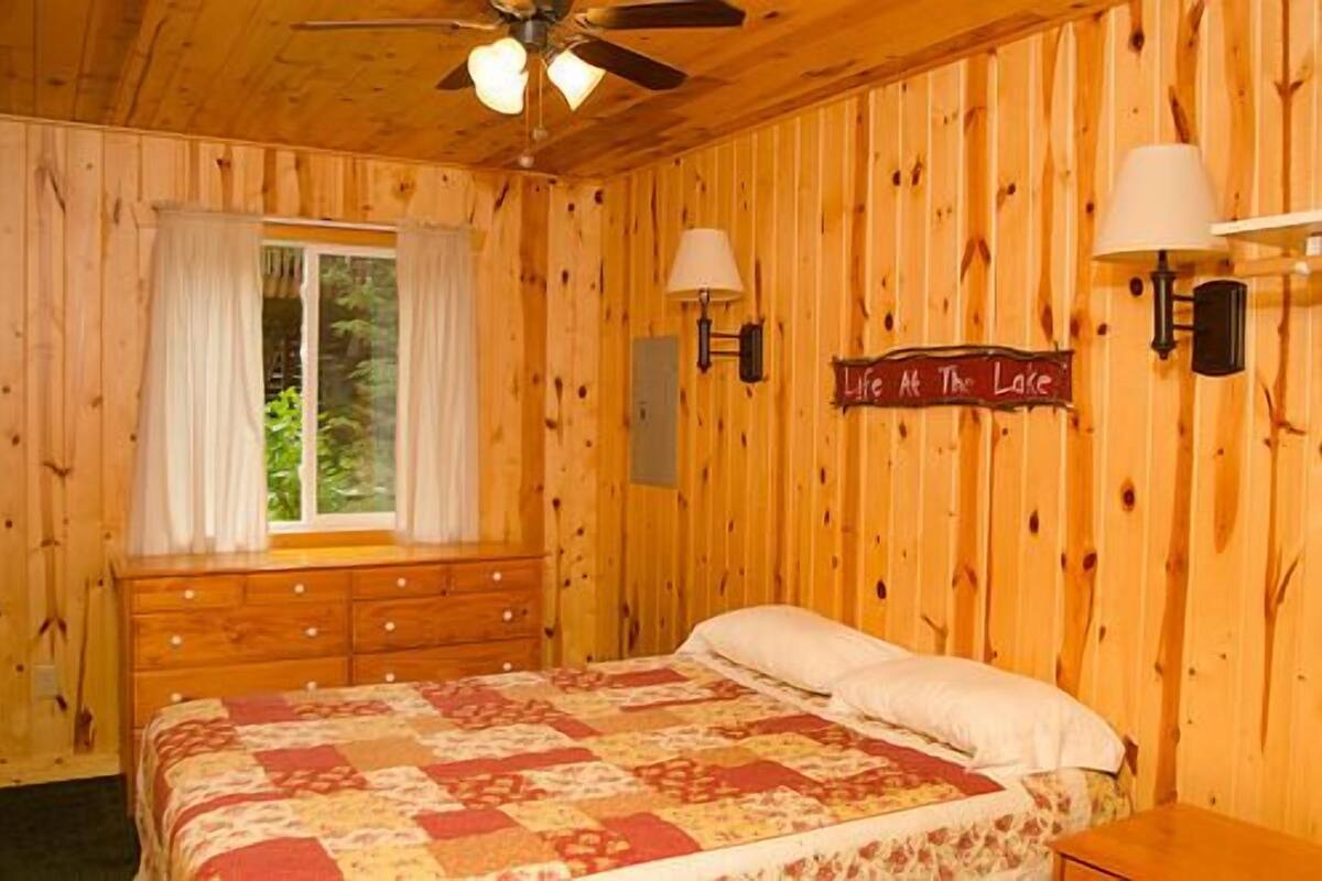 A wooden cabin bedroom with a bed, two wall-mounted lamps, a window with curtains, and a sign reading "Life at the Lake".