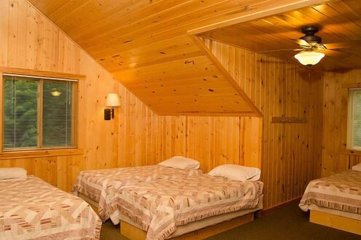 A room with wooden walls and a sloped ceiling. There are three beds with patterned bedding. A ceiling fan is visible, and a window with blinds is on the left wall.