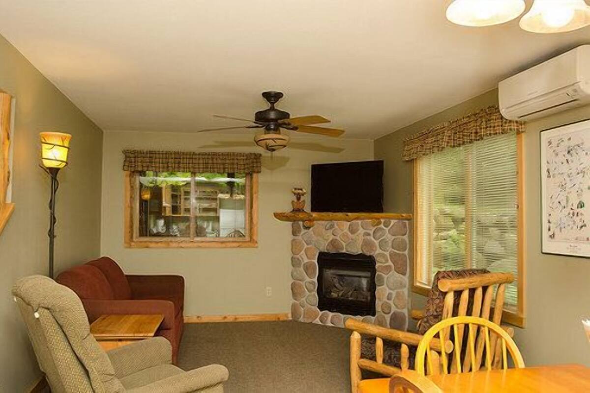 A living room with a stone fireplace, a ceiling fan, a television, and a window with blinds.