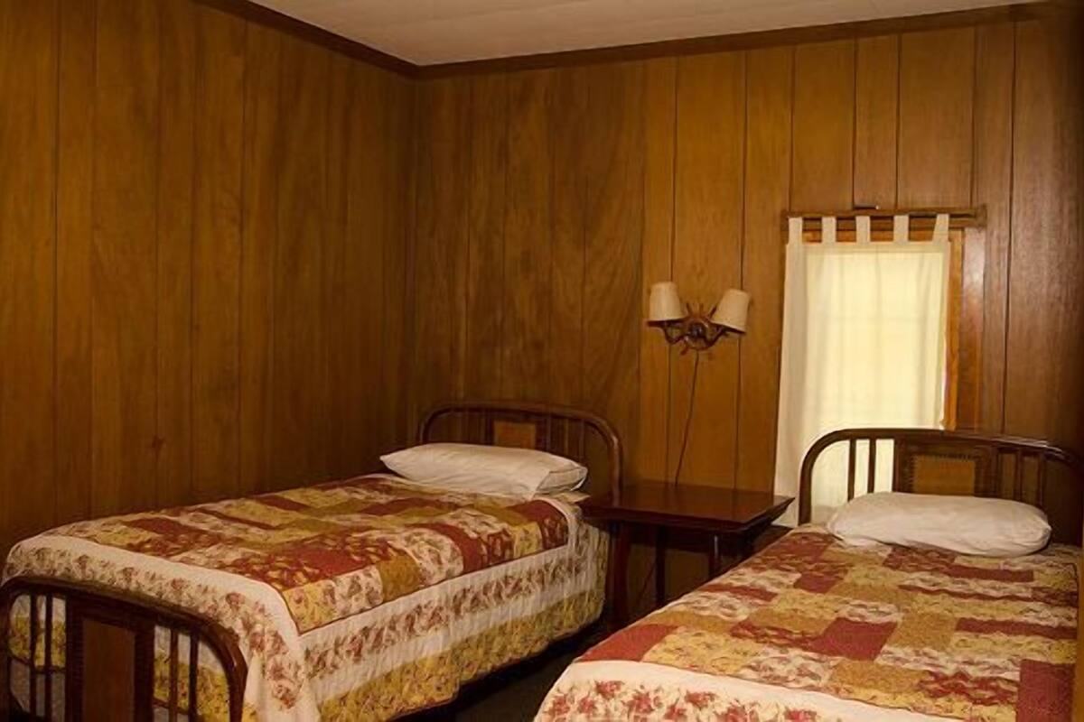 A room with two wooden beds, a small table, and a wall-mounted lamp.