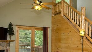 A spacious living area with a wooden staircase, a fireplace, and a large window overlooking a deck. - Woodland Beach Resort (Deerwood)