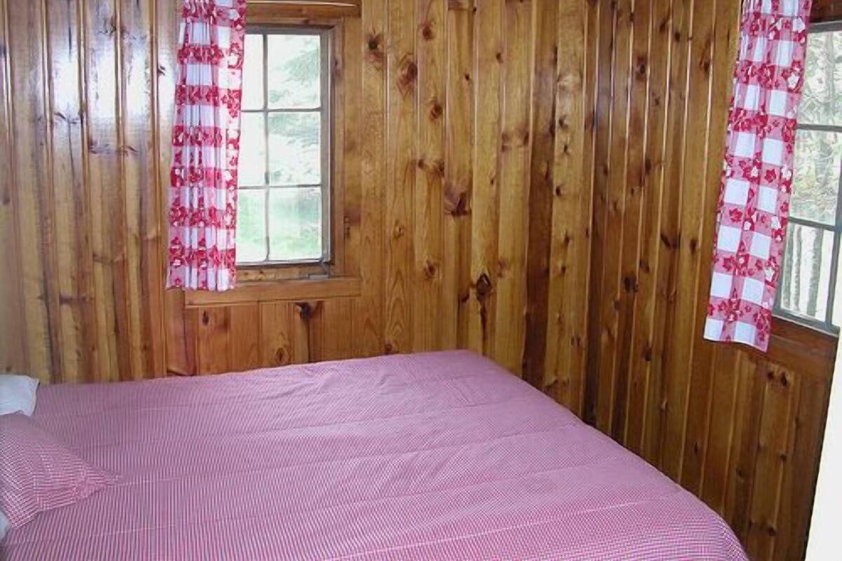 A room with wooden walls, a window with pink and white curtains, and a bed with a pink bedspread.
