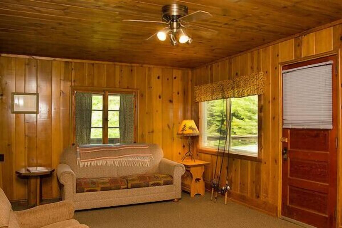 A cozy cabin interior with a wooden ceiling, a sofa, a small table, a lamp, and a window with a view of greenery.