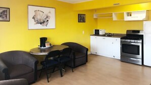 A compact kitchen with white cabinets, a microwave, and a stove. There is a dining area with two chairs and a small table. - Stampeder Motel (Williams Lake)
