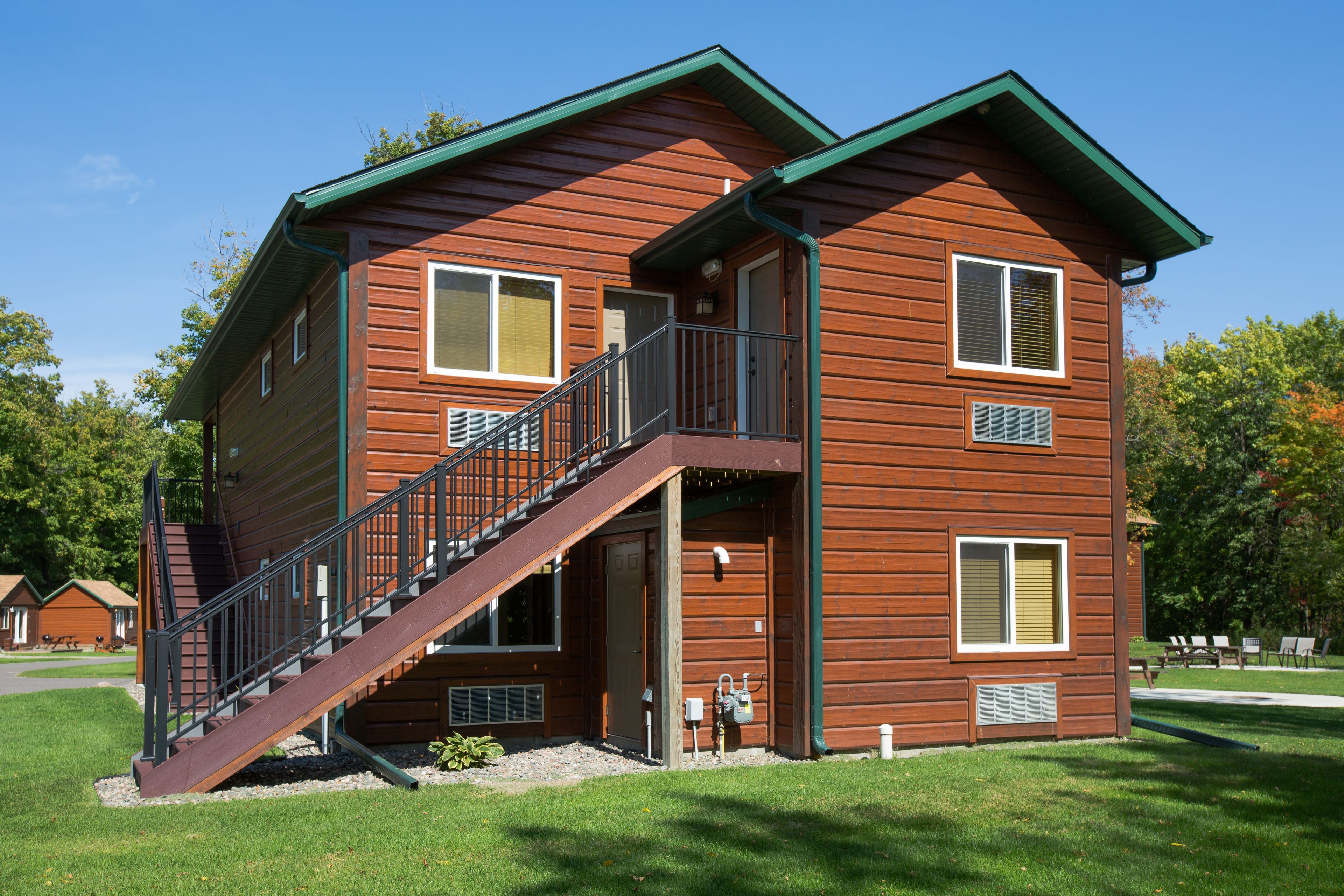 A two-story wooden cabin with a staircase leading to the upper floor, a small porch, and a clear view of the surrounding trees.