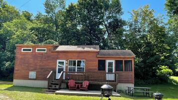 A wooden cabin with a deck, two red Adirondack chairs, a barbecue grill, and a picnic table.